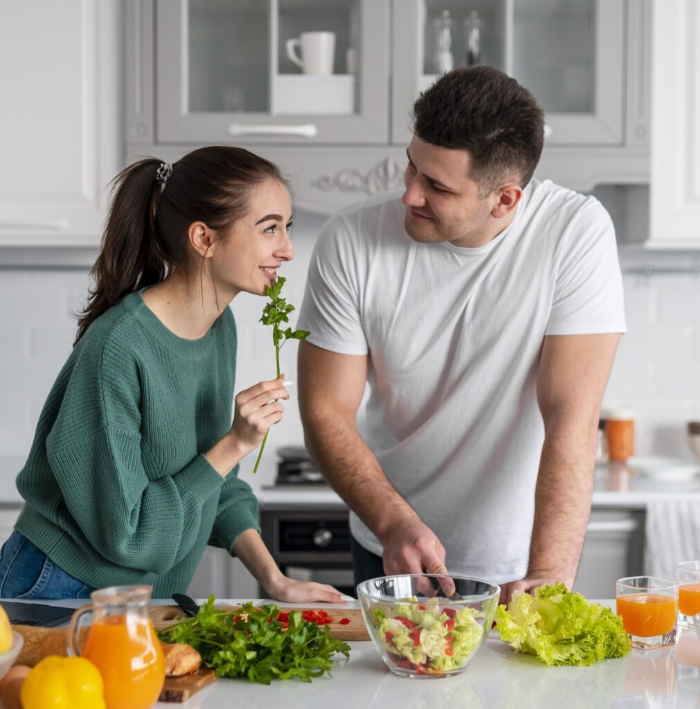 Casal preparando uma refeição saudável juntos na cozinha; a mulher segura uma folha de salsa enquanto o homem corta vegetais.