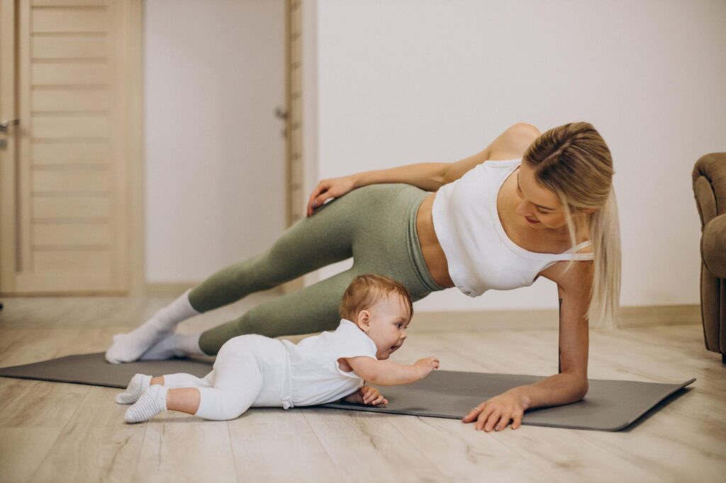 Mãe praticando exercício leve em casa com bebê ao lado sobre um tapete de yoga.