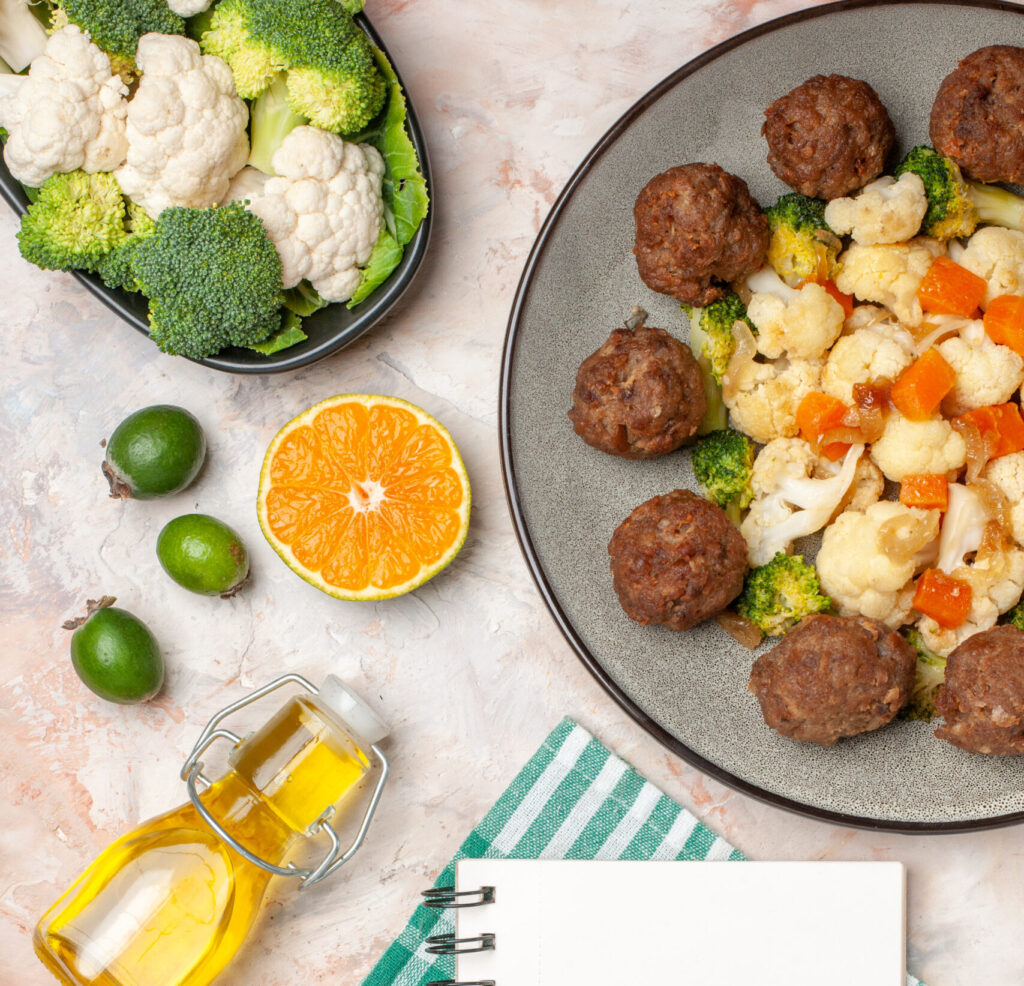Prato com almôndegas e vegetais cozidos (brócolis, couve-flor e cenoura), ao lado de uma bandeja com vegetais frescos, limões, uma laranja cortada, um frasco de azeite e um caderno em branco sobre um pano xadrez verde.